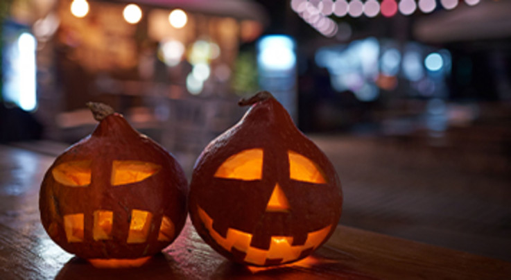 Zwei leuchtende Kürbislaternen mit geschnitzten Gesichtern stehen auf einem Holztisch, im Hintergrund verschwommene Lichter einer gemütlichen Abendstimmung.